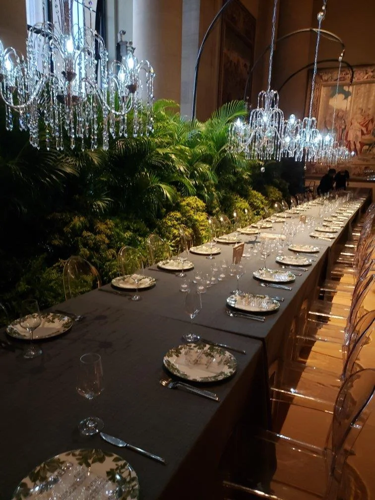 A long candlelit dining table beside layered palms, ferns, and hanging chandeliers.